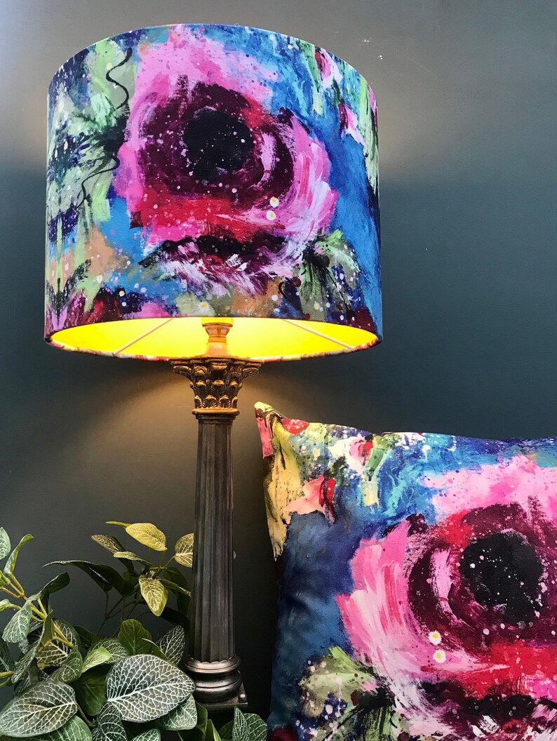 May include: A lampshade with a floral pattern in shades of pink, blue, and green. The lampshade is lit and the light shines through the fabric. The lampshade is on a black lamp base.