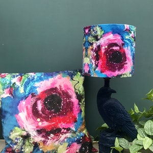 May include: A lamp with a blue, pink and white floral patterned lampshade and a black crow-shaped base. The lamp is next to a cushion with a similar floral pattern.