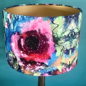 May include: A lampshade with a colourful abstract floral design. The lampshade is made of fabric and has a gold trim. The design features pink, blue, green, and black colours.