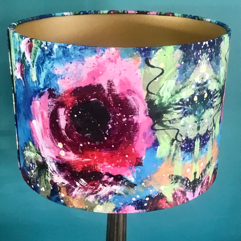 May include: A lampshade with a colourful abstract floral design. The lampshade is made of fabric and has a gold trim. The design features pink, blue, green, and black colours.