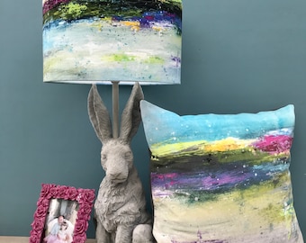 May include: A lamp with a colourful abstract design lampshade, a grey rabbit sculpture, and a cushion with a similar abstract design. The cushion is on a table with a pink picture frame.
