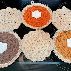 May include: A collection of six handmade crochet pot holders in the shape of pies. The pot holders feature various colors, including brown, tan, and orange, with white accents resembling whipped cream. Each has a scalloped edge.