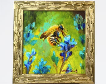 Può includere: Un piccolo dipinto di un'ape su un fiore. L'ape è gialla e nera, e il fiore è blu e viola. Il dipinto è incorniciato in oro ed è su un piccolo cavalletto.