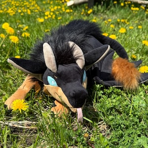 May include: A plush creature with black fur, tan ears, and blue eyes rests in a field of green grass and yellow dandelions. The creature has a long tail with a fluffy, orange tuft and a pink tongue.