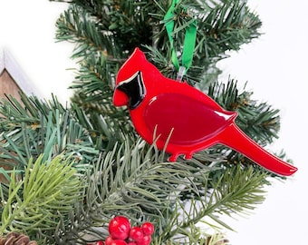 May include: A glossy red cardinal ornament with a black accent on its face hangs from a green ribbon on a Christmas tree. The bird-shaped decoration is a festive holiday item.