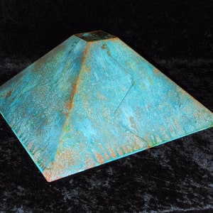 Puede incluir: Una pirámide cuadrada de color turquesa con una superficie texturizada. La pirámide está hecha de metal y tiene un aspecto ligeramente envejecido.