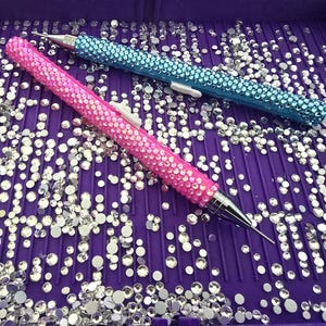 May include: Two rhinestone-covered craft tools, one pink and one blue, rest on a purple tray filled with scattered rhinestones. The tools have silver tips and white accents. The pink tool is on the bottom left, and the blue tool is on the top right.