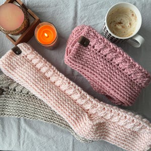 May include: Three crocheted headbands in light pink, dusty rose, and beige, each featuring a textured cable knit pattern and a small brown leather-like tag with a metal rivet. The headbands are arranged alongside a lit orange candle and a mug.