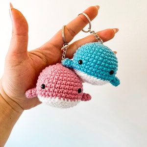 May include: Two small, round, crocheted whale keychains, one in light pink and one in bright blue, each featuring a white underbelly, tiny side fins, and small black bead eyes, attached to silver-toned metal key rings with short link chains.