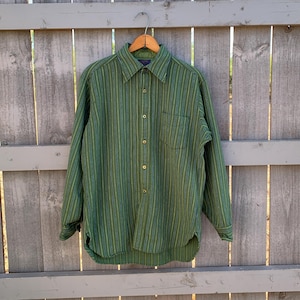 May include: A long-sleeve button-down shirt featuring a vertical striped pattern in shades of olive green, forest green, and navy blue. The shirt includes a pointed collar, a single chest pocket on the left side, and light-colored buttons down the front.