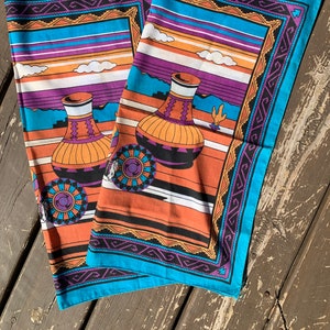 May include: Two turquoise blue fabric squares with a Southwestern design. The fabric features a repeating pattern of orange, purple, and black stripes, with a stylized image of two pottery jars in the center. The border of the fabric is a repeating pattern of black, purple, and orange.
