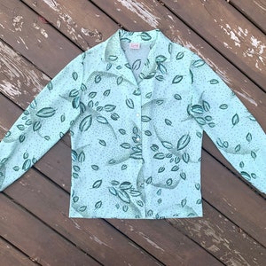 May include: A light blue long-sleeved shirt with a green leaf pattern. The shirt has a button-up front and a collared neckline. The label on the shirt reads "Gerry".