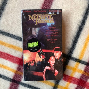 May include: A VHS tape for the horror movie Needful Things, based on the Stephen King novel. The cover features a dark, ominous scene with a man and woman in the foreground, and a house in the background. The title of the movie is in large, white letters on a black background. The text "The town of Castle Rock just made a deal with the devil... Now it's time to pay." is printed in white on a black background.