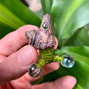 Puede incluir: Una pequeña escultura de vidrio soplado a mano con una cabeza marrón texturizada y piernas verdes rayadas. La escultura tiene tres orbes redondos de vidrio transparente en los extremos de las piernas.