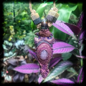 May include: A purple and copper arrowhead pendant with a pink gemstone in the center, hanging from a brown cord with two layered stone beads.