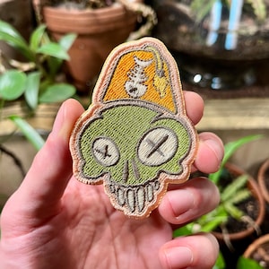 May include: Green skull embroidered patch with a yellow fez hat with a fishbone design. The patch has a stitched outline and is held in a hand.