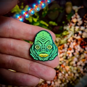 May include: Green enamel pin featuring a cartoon illustration of the Creature from the Black Lagoon. The pin has a yellow mouth and eyes.