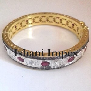 Puede incluir: Pulsera de oro con una banda de plata engastada con piedras preciosas rojas y blancas. La pulsera tiene un diseño decorativo recortado en la parte superior y las palabras "Ishani Impex" están grabadas en la banda de plata.