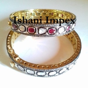 May include: A pair of gold bangles with a silver band set with small diamonds and red rubies. The bangles are stacked on top of each other and the top bangle has the text "Ishani Impex" on it.