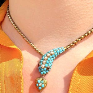 May include: A gold snake necklace featuring a head encrusted with small turquoise beads and white pearls, with two dark red stone eyes. A small gold heart-shaped pendant hangs from the snake's mouth, decorated with turquoise beads and a central pearl.