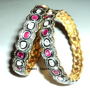 Pode incluir: Dois braceletes dourados com um design de borda recortada e pedras ovais vermelhas e transparentes alternadas, incrustadas em metal preto e prateado. O texto Ishani Impex está impresso em letras pretas na superfície dos braceletes.