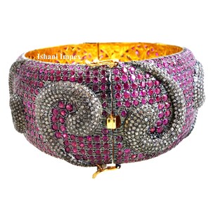 May include: A wide hinged bangle bracelet featuring a dense pavé setting of vibrant pink rubies and dark grey diamonds arranged in swirling patterns. The interior is gold-toned, and the text Ishani Impex is visible on the inner band surface.