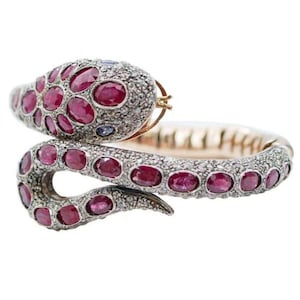 May include: A silver and gold snake bracelet with red rubies and diamonds. The snake is coiled and the head is raised. The bracelet is made of metal and has a hinged clasp.