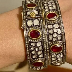Könnte beinhalten: Zwei silberne Armreifen mit roten Rubin-Steinen und Pavé-Diamanten. Die Armreifen sind auf dem Handgelenk einer Person übereinander gelegt.