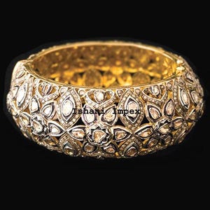 Könnte beinhalten: Ein goldfarbenes Armreif-Armband mit einem aufwendigen floralen und geometrischen Design. Das Armband ist mit zahlreichen kleinen, klaren Edelsteinen besetzt, die einen funkelnden Effekt erzeugen. Der Text "Ishani Impex" ist sichtbar.