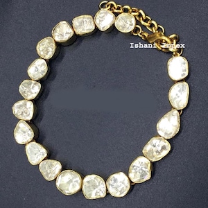 Puede incluir: Una pulsera de tono dorado con una serie de piedras transparentes irregulares y facetadas engastadas en metal. La pulsera incluye una cadena de extensión dorada con un cierre de mosquetón y el texto Ishani impex impreso cerca del cierre.