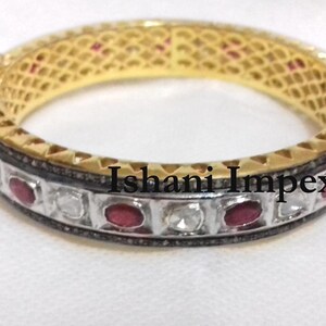 Könnte beinhalten: Ein goldenes Armreif-Armband mit einem silbernen Band, das mit kleinen weißen Diamanten und roten Rubinen besetzt ist. Das Armband ist mit dem Text "Ishani Impex" graviert.