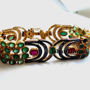 May include: A gold bracelet with emerald green and ruby red gemstones set in a black metal design. The bracelet has a clasp closure.