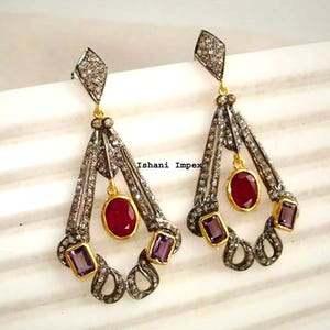 May include: A pair of chandelier earrings featuring a diamond-encrusted frame with a central oval red gemstone and two rectangular purple gemstones. The metalwork is dark with gold accents. The text Ishani Impex is visible in the center.