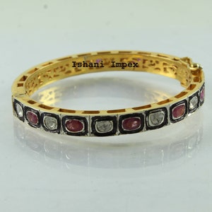 May include: Gold bangle bracelet with a black enamel border and a row of alternating red and white gemstones. The bracelet is engraved with the text "Ishani Impex".