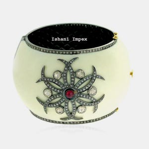 Könnte beinhalten: Cremefarbenes Armband mit floralem Design. Das Armband hat einen zentralen roten Edelstein, umgeben von einem silberfarbenen Blumenmuster mit klaren Edelsteinen. Der Text "Ishani Impex" ist auf der Innenseite des Armbands zu sehen.