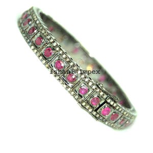 May include: A hinged bangle bracelet featuring a dark oxidized metal finish with a row of round, vibrant pink gemstones set in square frames, accented by small white pavé stones. The text Ishani Impex is visible on the bracelet surface.