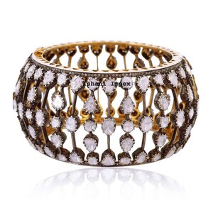 Puede incluir: Una pulsera ancha y ornamentada con un interior dorado y un exterior de metal oscuro. La pulsera está incrustada con numerosas piedras preciosas claras de forma irregular. El texto "ishani Impex" es visible.