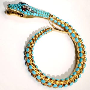 May include: A gold snake bracelet featuring a head and body covered in small, round, bright blue turquoise cabochons. The snake head has a red stone eye, clear accent stones, and an open mouth. The text Ishahi impex is visible on the body.