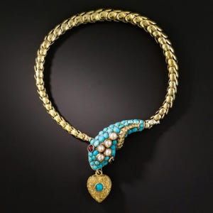 Op de afbeelding: Een goudkleurige slangvormige ketting met een flexibele schakelketting in schubpatroon. De kop is ingelegd met kleine turkoois stenen en witte parels, met een oog van een rode steen. Een getextureerde gouden hartvormige hanger met turkoois hangt onderaan.
