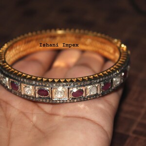 Op de afbeelding: Gouden armband met afwisselend heldere en rode edelstenen. De armband heeft een getextureerde gouden band en een donkere metalen zetting voor de edelstenen. De tekst "Ishani Impex" is zichtbaar.