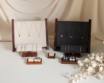 Puede incluir: Expositores de joyería con collares, pulseras y pendientes. Los expositores tienen marcos de madera y están cubiertos de tela blanca o negra. Las joyas son doradas y se exhiben en los expositores. El fondo es de color neutro.