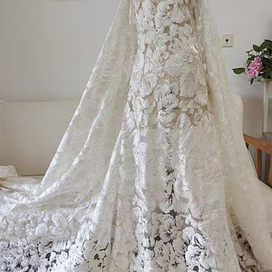 May include: White floral lace fabric with a delicate, intricate pattern. The lace is sheer and has a soft, flowing texture. The fabric is perfect for creating elegant and romantic garments.