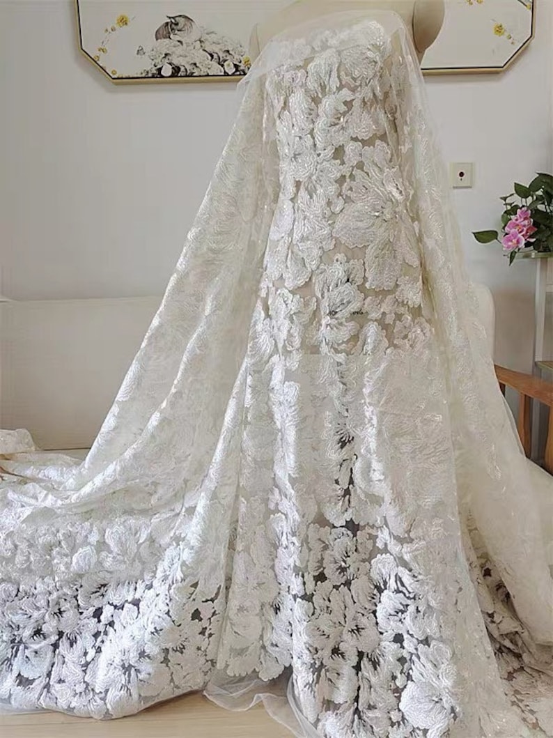 May include: White floral lace fabric with a delicate, intricate pattern. The lace is sheer and has a soft, flowing texture. The fabric is perfect for creating elegant and romantic garments.