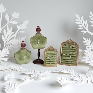 Könnte beinhalten: Ein olivgrünes Schmuck-Display-Set aus Samt mit einer Halskettenbüste und zwei verzierten Ringhaltern. Die Ringhalter haben einen goldenen Rahmen und eine grüne Samtinnenseite. Weiße Papierblattausschnitte sind um die Präsentation verstreut.