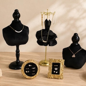 May include: Two black velvet jewelry busts, a gold-toned metal dress-shaped necklace stand, and two ornate gold-toned frames with black velvet inserts for rings. The jewelry displays hold various necklaces, earrings, and rings on display.
