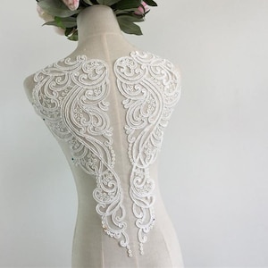 May include: White lace applique with a delicate floral design. The applique is shaped like two wings and has a scalloped edge.
