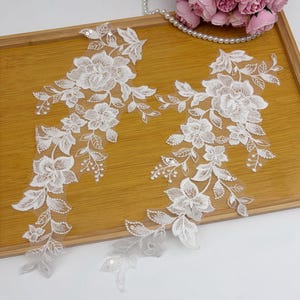 Può includere: Due applicazioni in pizzo floreale bianco con intricati disegni di fiori e foglie. Le applicazioni sono impreziosite da paillettes e sono poste su un vassoio di legno marrone chiaro. Una collana di perle e fiori rosa sono sullo sfondo.