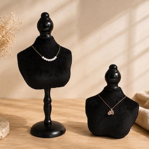 May include: Two black velvet jewelry display busts on stands. The taller bust features a delicate gold chain necklace with seven small white pearls. The shorter bust displays a gold chain necklace with a small circular pendant featuring a star design.