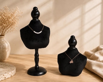 May include: Two black velvet jewelry display busts on stands. The taller bust features a delicate gold chain necklace with seven small white pearls. The shorter bust displays a gold chain necklace with a small circular pendant featuring a star design.