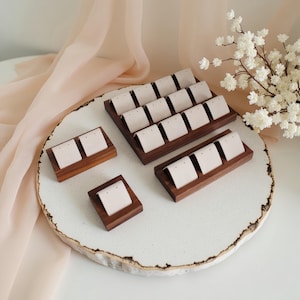 Könnte beinhalten: Ohrring-Präsentationsständer aus Holz mit cremefarbenen Einsätzen, in verschiedenen Größen und Formen, angeordnet auf einem weißen Tablett mit organischem Rand. Das Holz hat eine dunkelbraune Oberfläche. Ein weicher, pfirsichfarbener Stoff und weiße Blumen sind im Hintergrund.